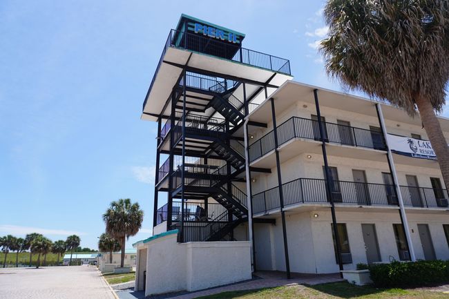 Modern building with exterior stair tower, white balconies, and a palm tree beside a sunny walkway