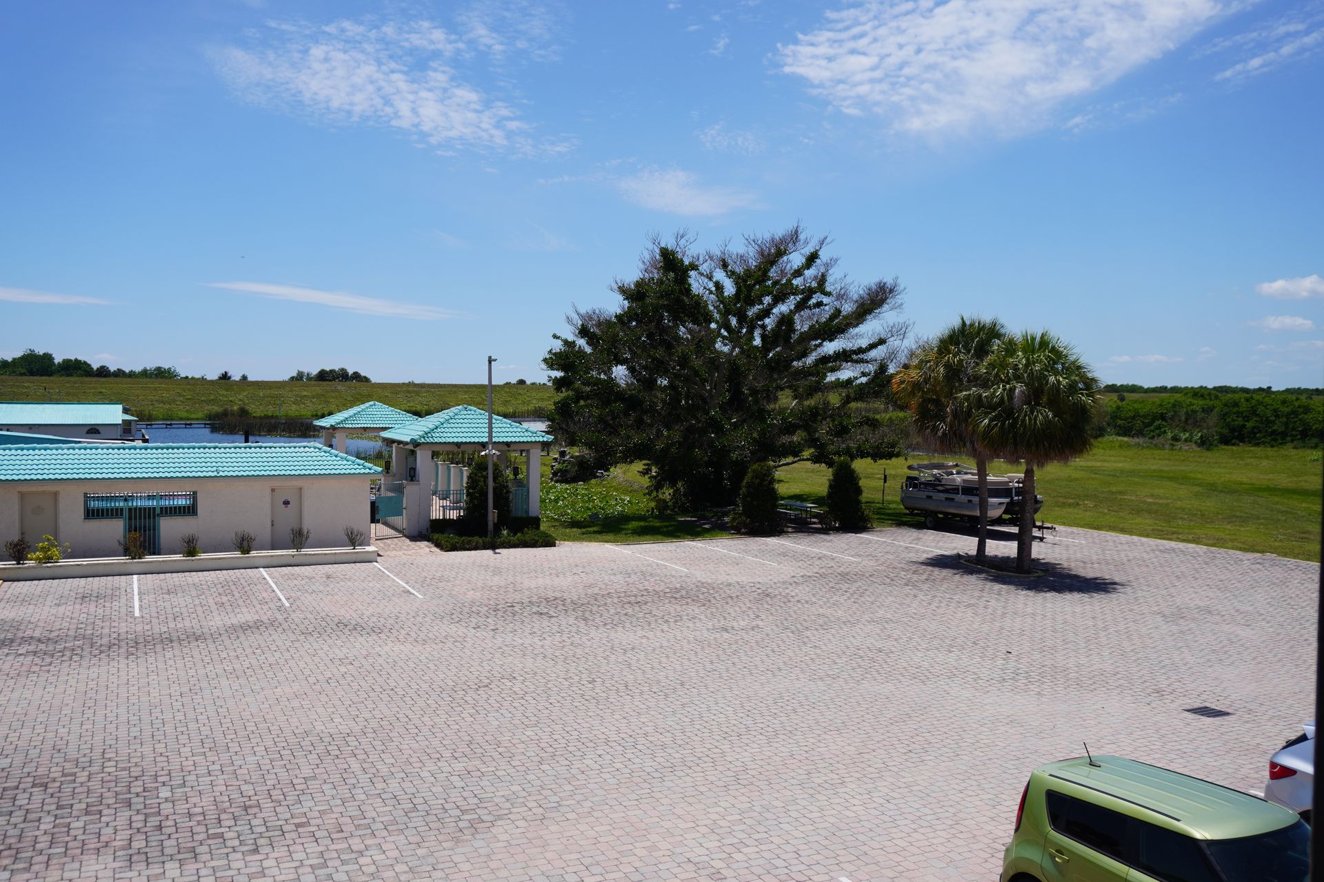 Gravel parking area beside a small building with teal umbrellas, trees, and open green field under a blue sky