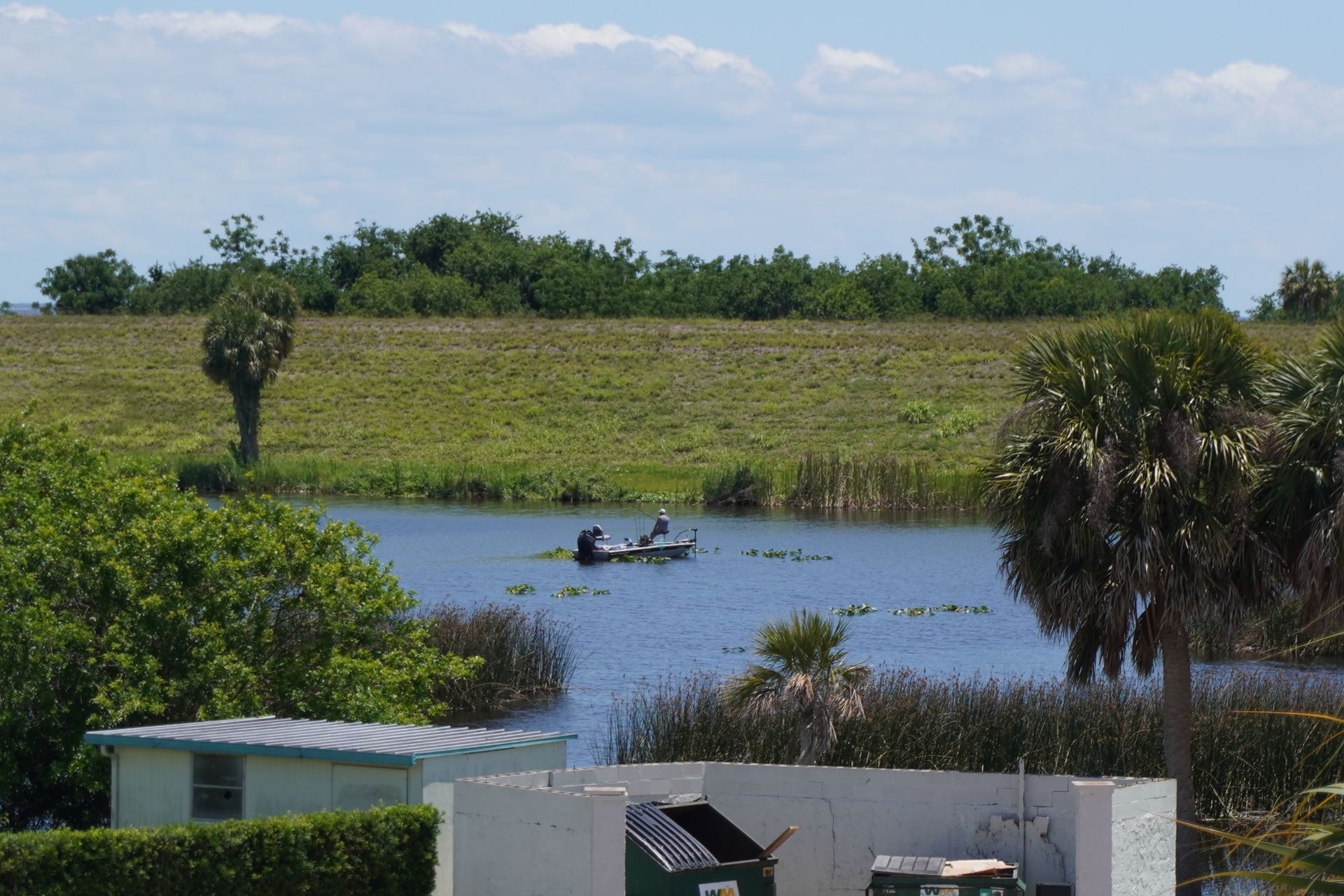 Small boat on a blue lagoon with grassy banks, palm trees, and buildings in the foreground under a cloudy sky