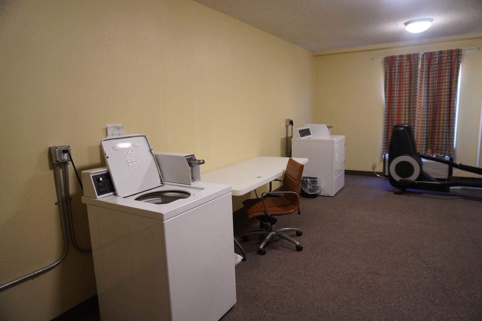 Small office with desks, computer equipment, a chair, and a treadmill near a window with curtains.