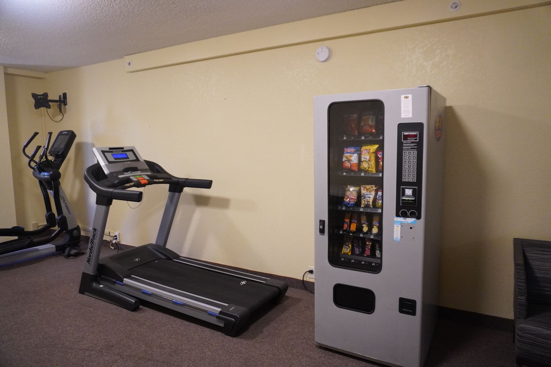 Gym room with treadmill, elliptical, and snack vending machine along a beige wall.