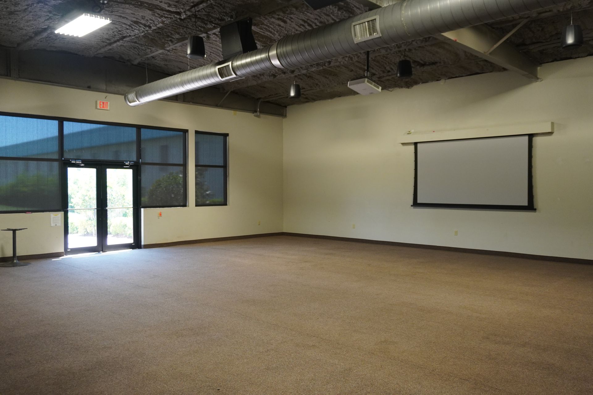 Empty carpeted room with beige walls, large windows, and a projector screen on the right.