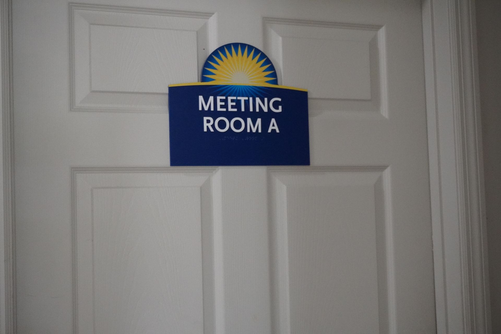 Blue “Meeting Room A” sign on a white door with a sunburst graphic above it
