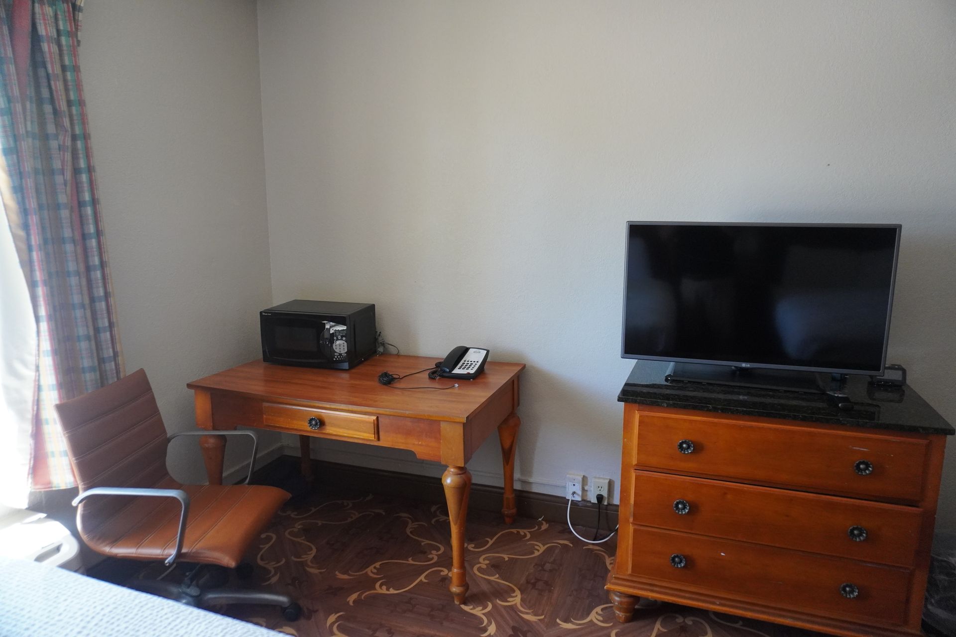 Small living room with wooden desk, TV stand, chair, and patterned carpet by a window