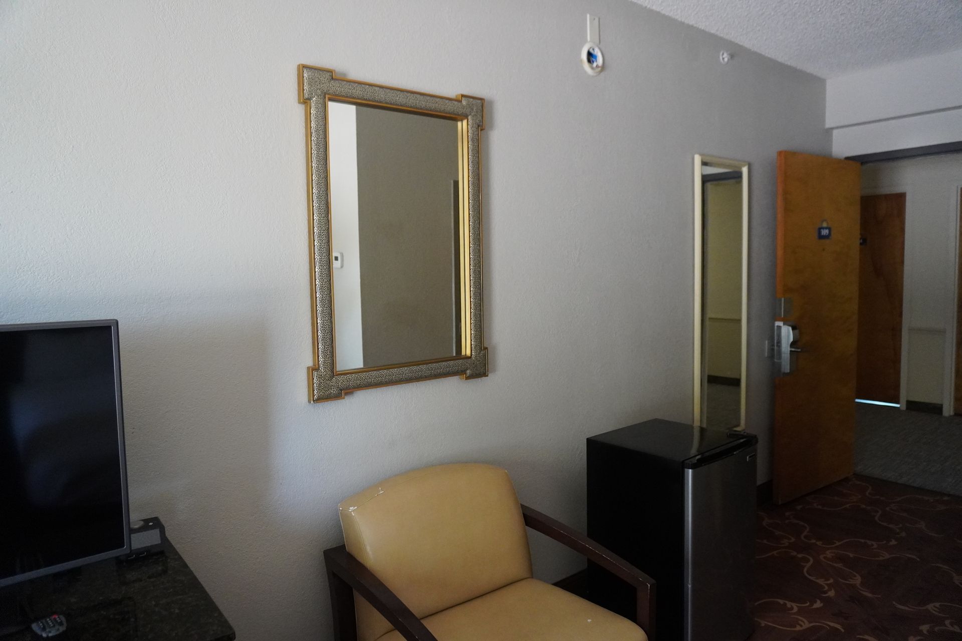 Hotel room interior with chair, wall mirror, TV edge, and open door to hallway