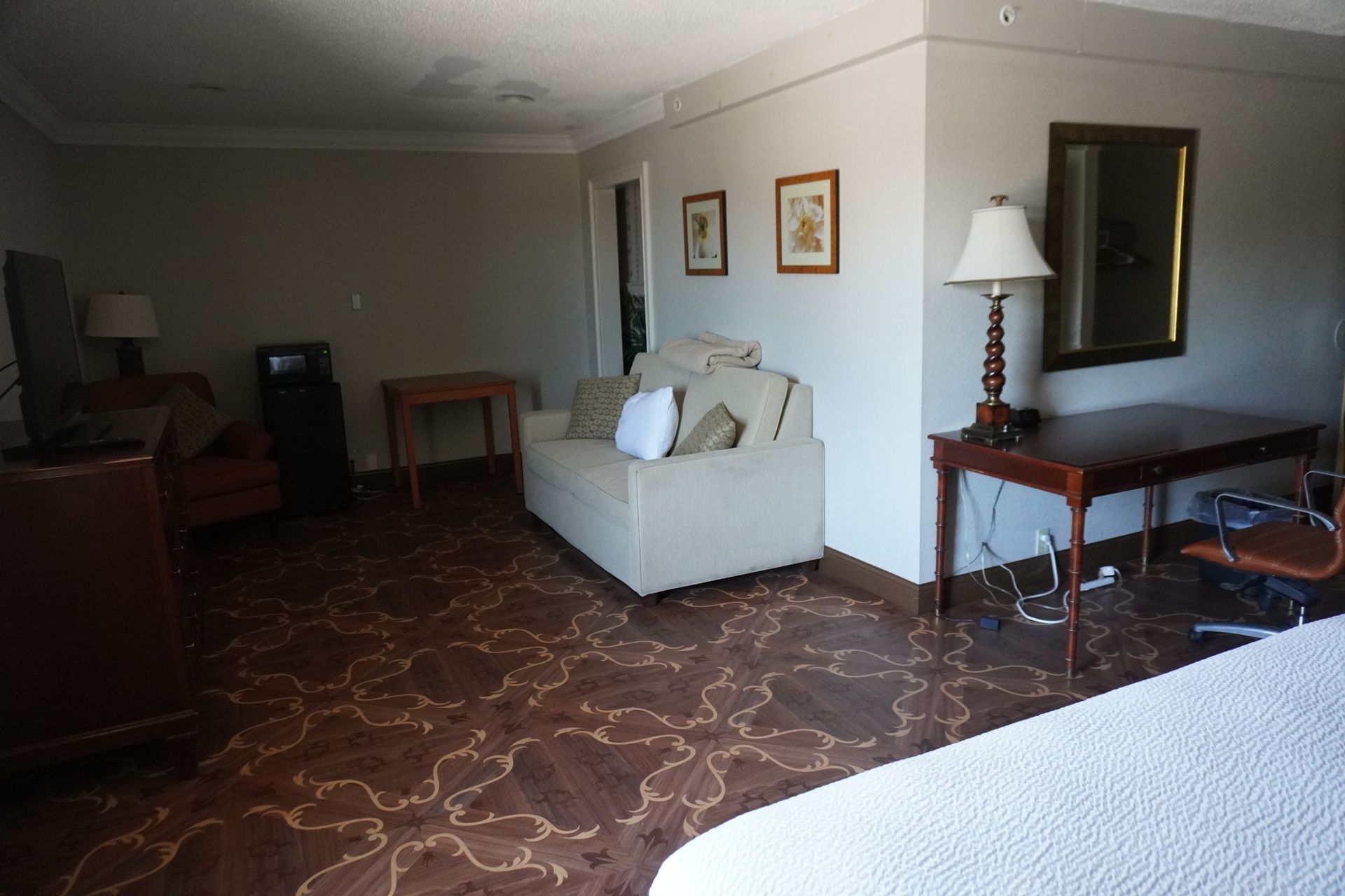 Spartan hotel room with sofa, table, lamp, wall art, and patterned carpet.