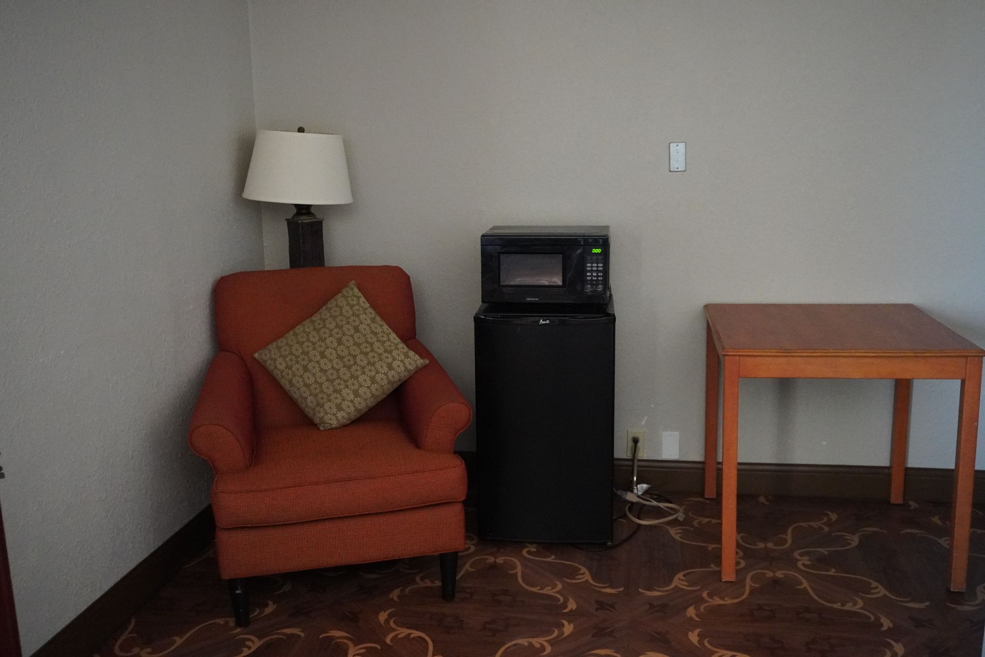 Small room with red armchair, lamp, microwave on mini fridge, and wooden table.
