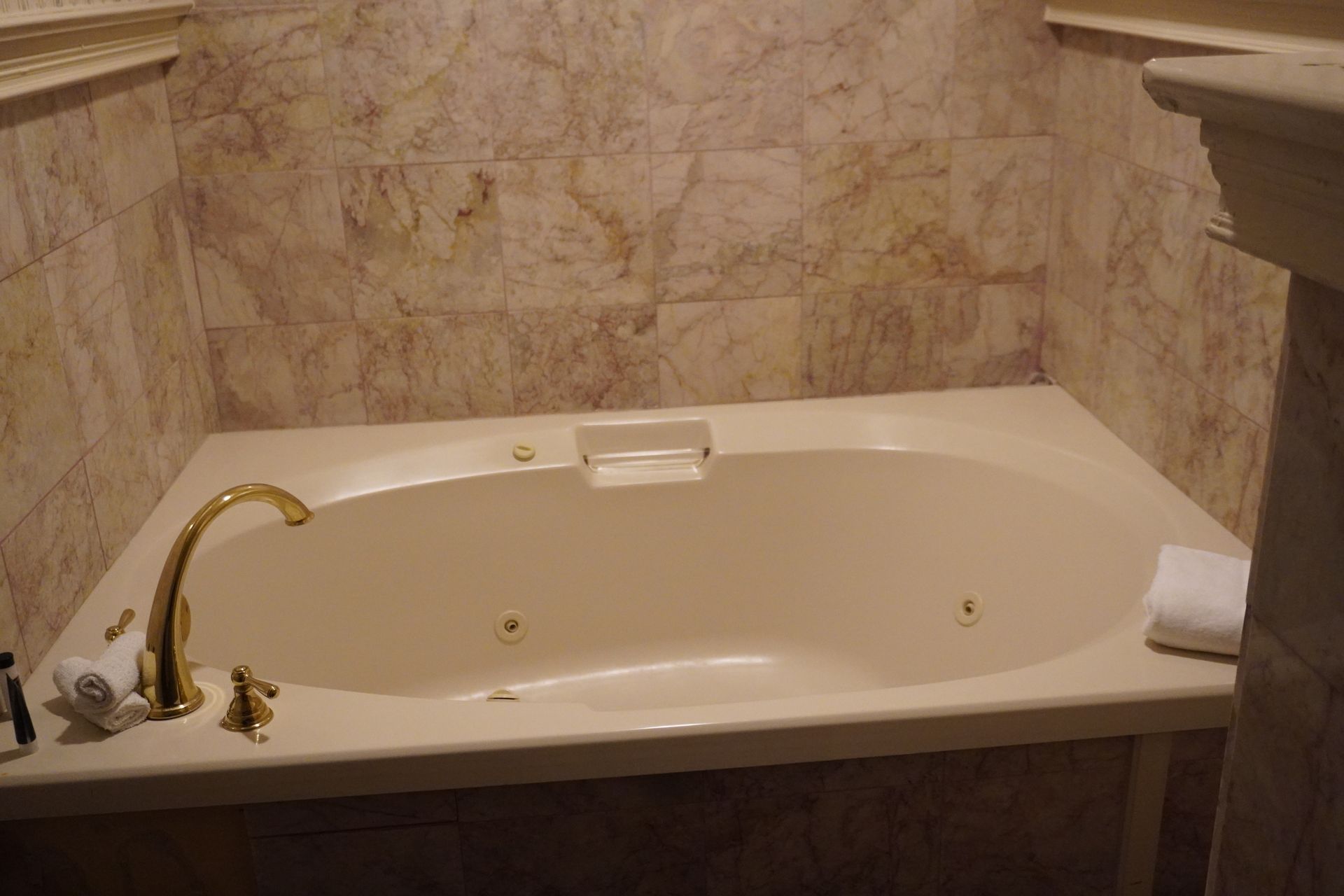 Corner bathtub with gold faucet and beige marble walls in a bathroom