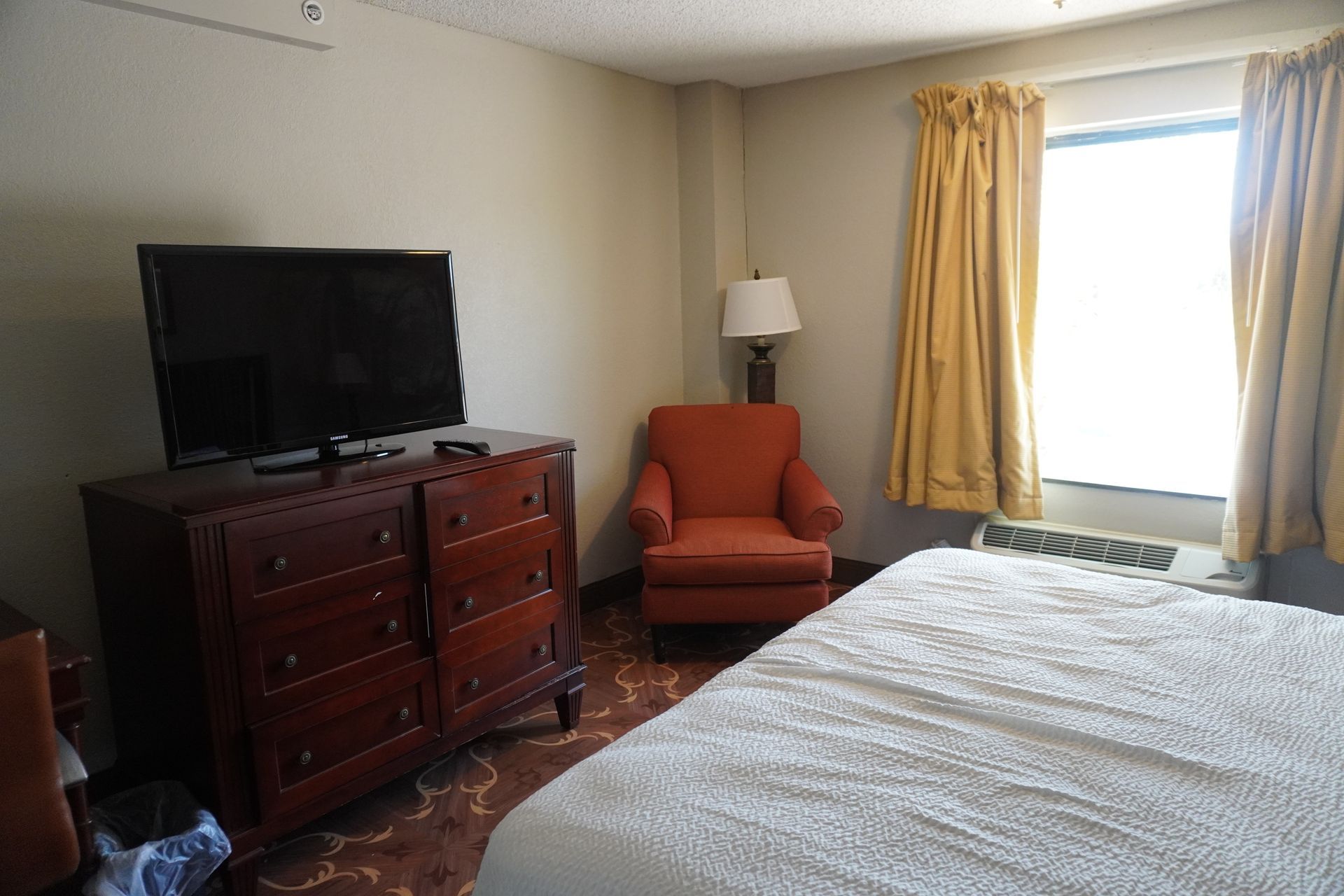 Bedroom with a bed, dresser and TV, orange chair, floor lamp, and bright window with curtains.