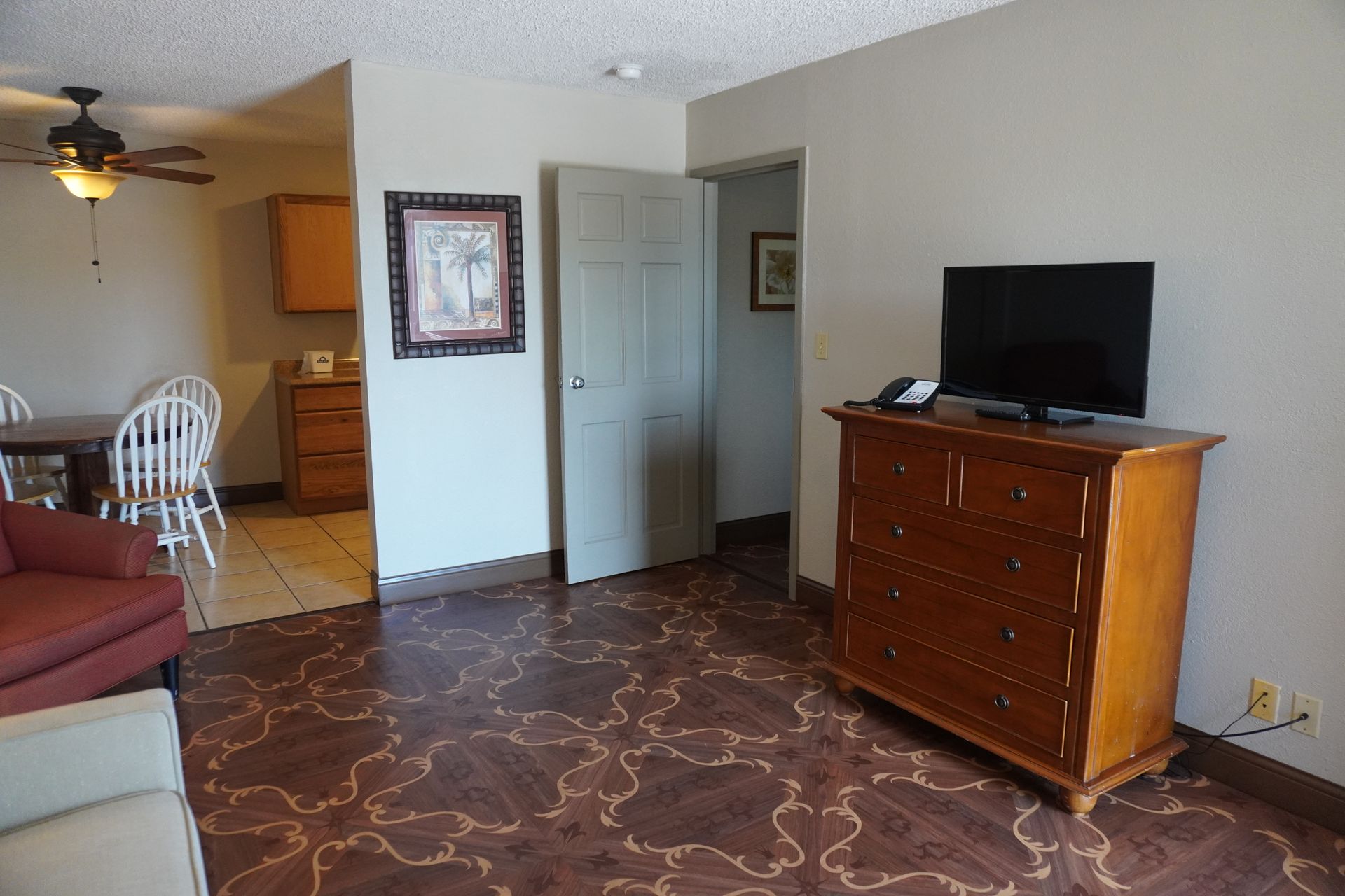 Living room with patterned carpet, wooden dresser and TV, opening to a dining area and kitchen.