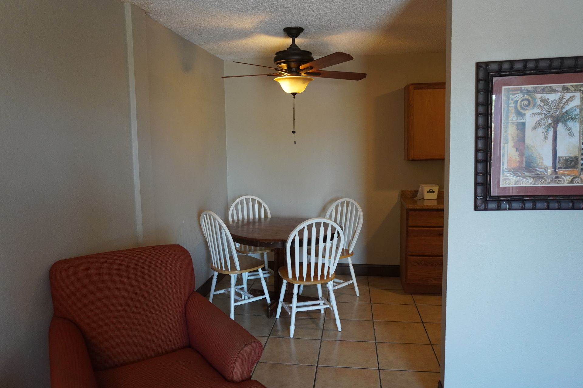 Dining area with round table, white chairs, ceiling fan, and red armchair in a beige room.