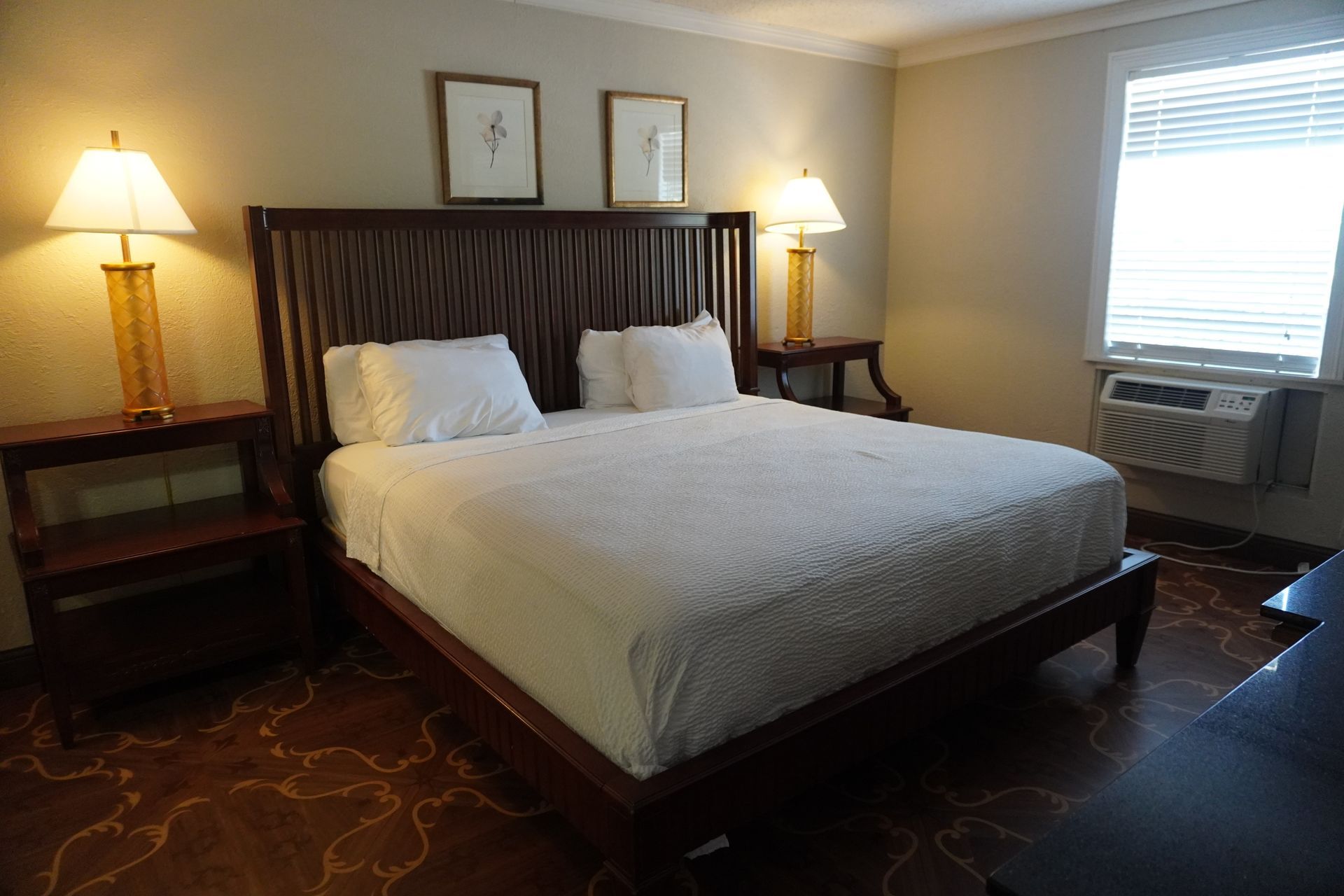 Hotel room with a large bed, two bedside lamps, framed art, and a window air conditioner.