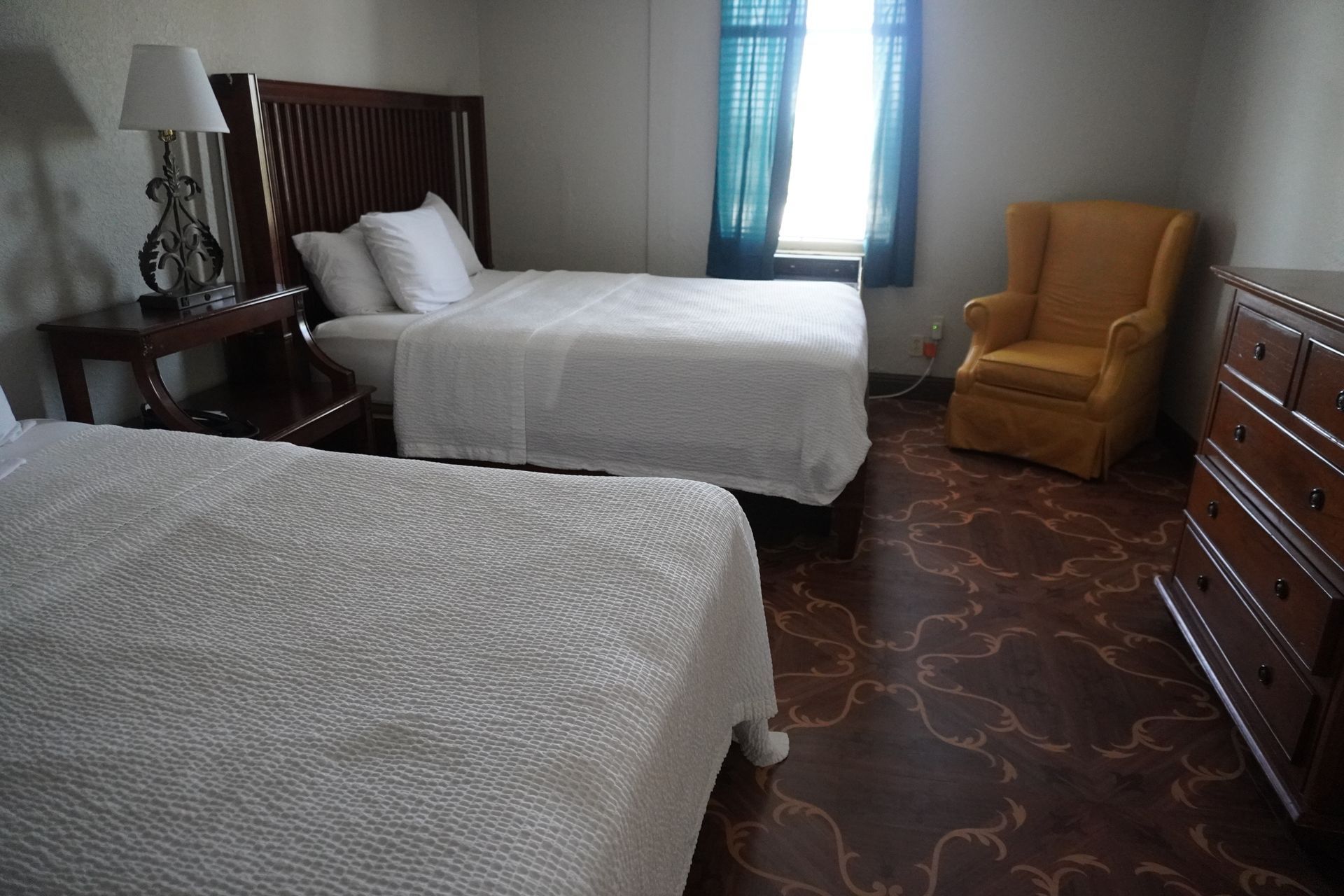 Small bedroom with two beds, a window, a yellow chair, and a dresser on a dark wood floor.