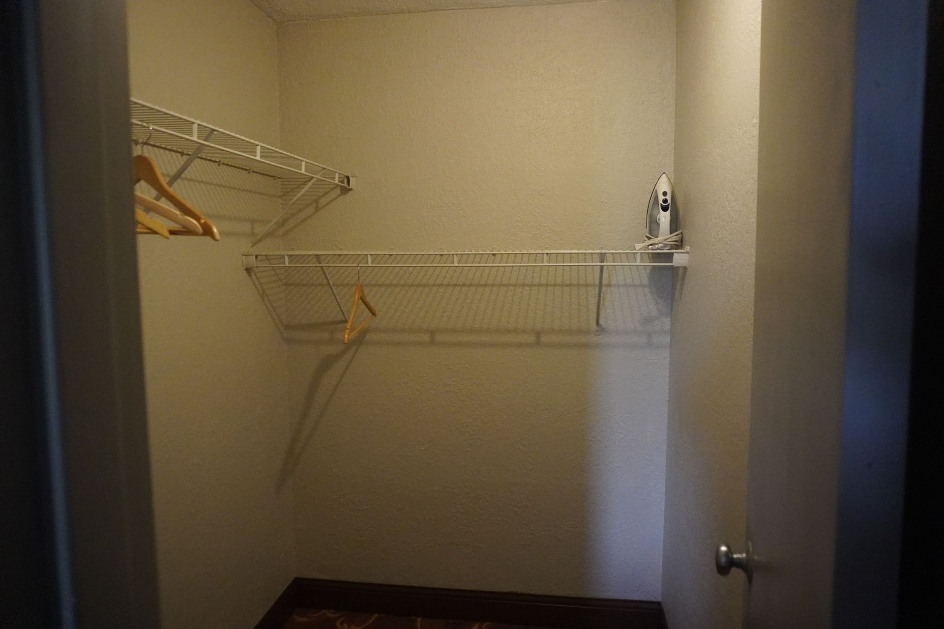Empty walk-in closet with white wire shelves and hanging rods, beige walls, and a dark carpeted floor