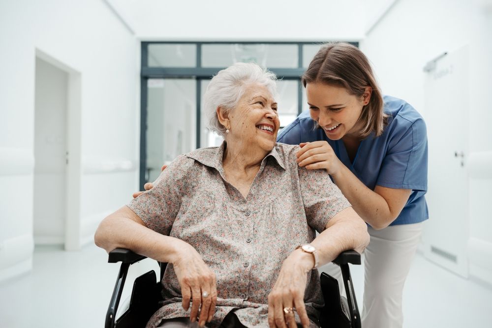 Caregiver smiling at an elderly woman in a wheelchair in a bright hallway