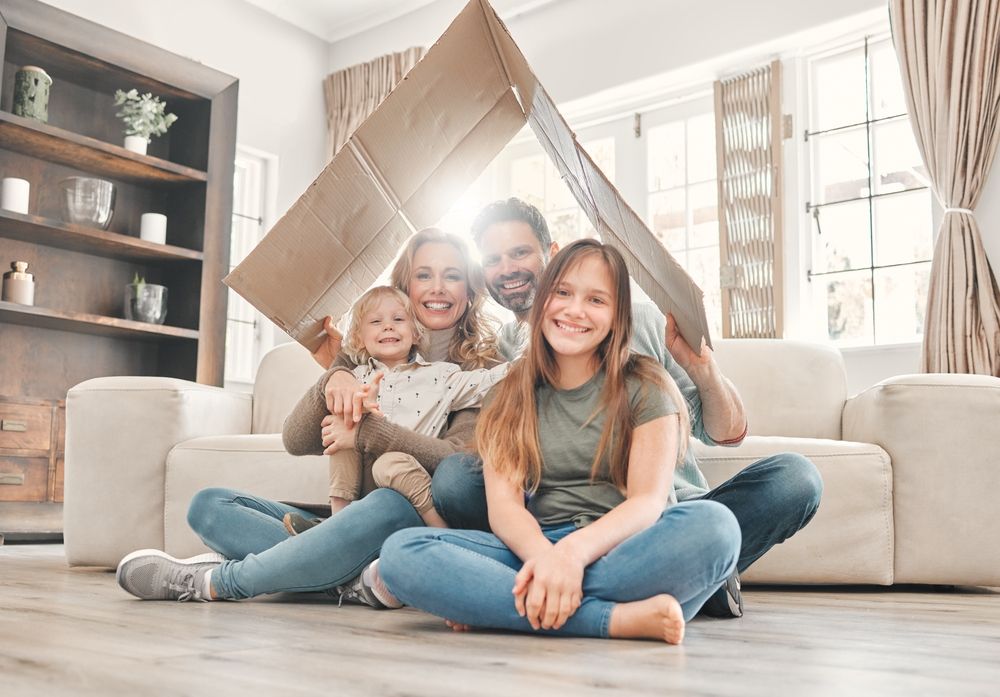 Three smiling children sit on the floor in a bright living room with a couch and shelves behind them.
