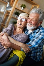 Smiling couple cuddling on a couch in a cozy living room, with shelves and plants in the background.