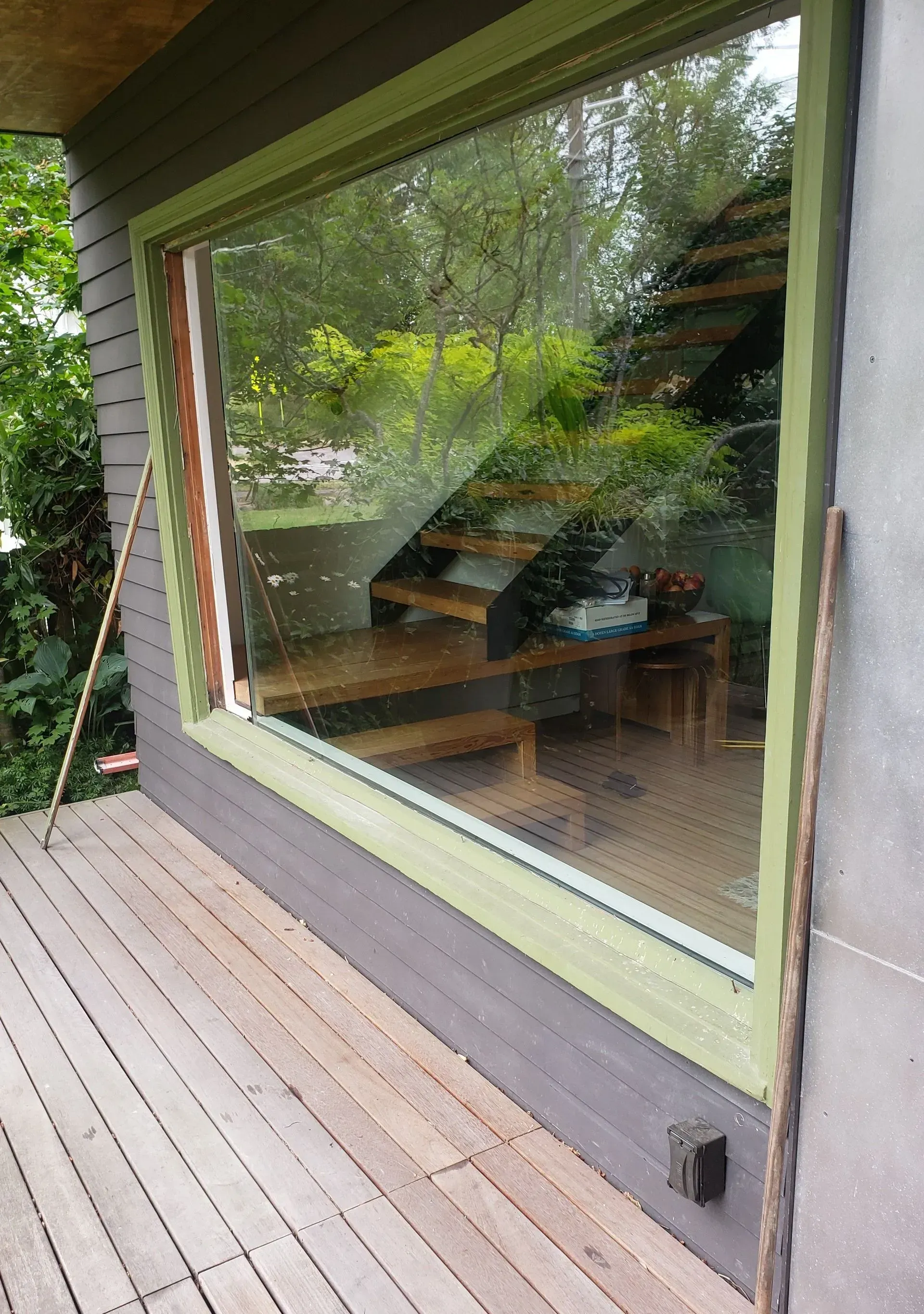 A large window with a light green frame overlooks a deck, revealing a glimpse of interior stairs and a wooden table.