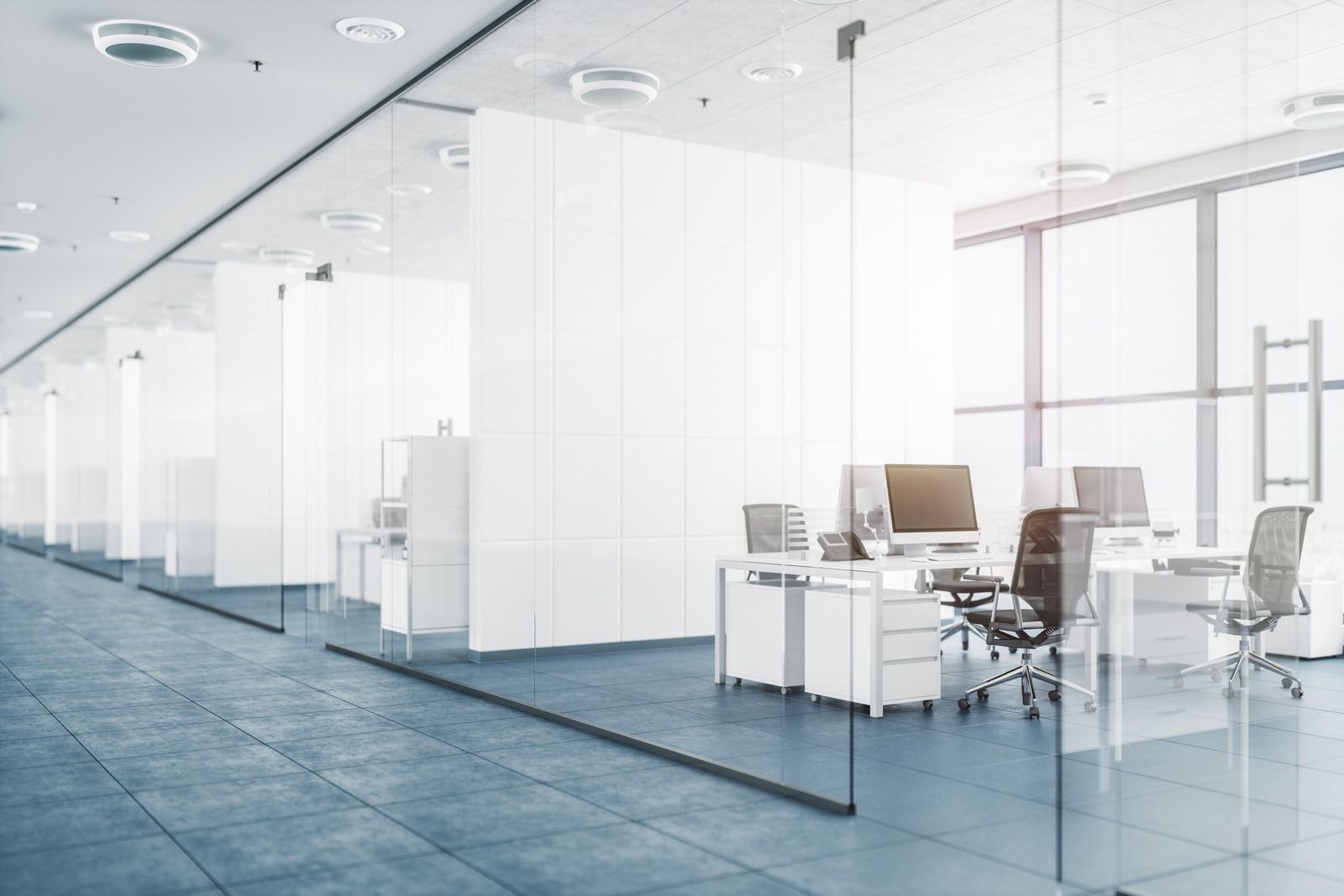 A modern, bright office interior with glass-walled cubicles, white desks, rolling chairs, and large windows.