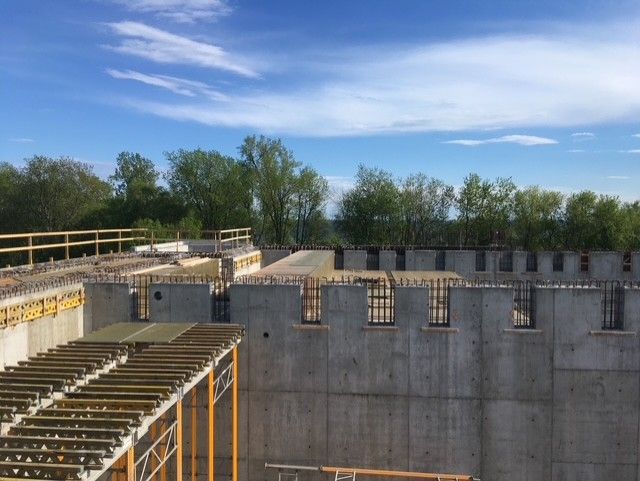 Concrete building under construction with exposed walls, wooden scaffolding, and trees under a blue sky