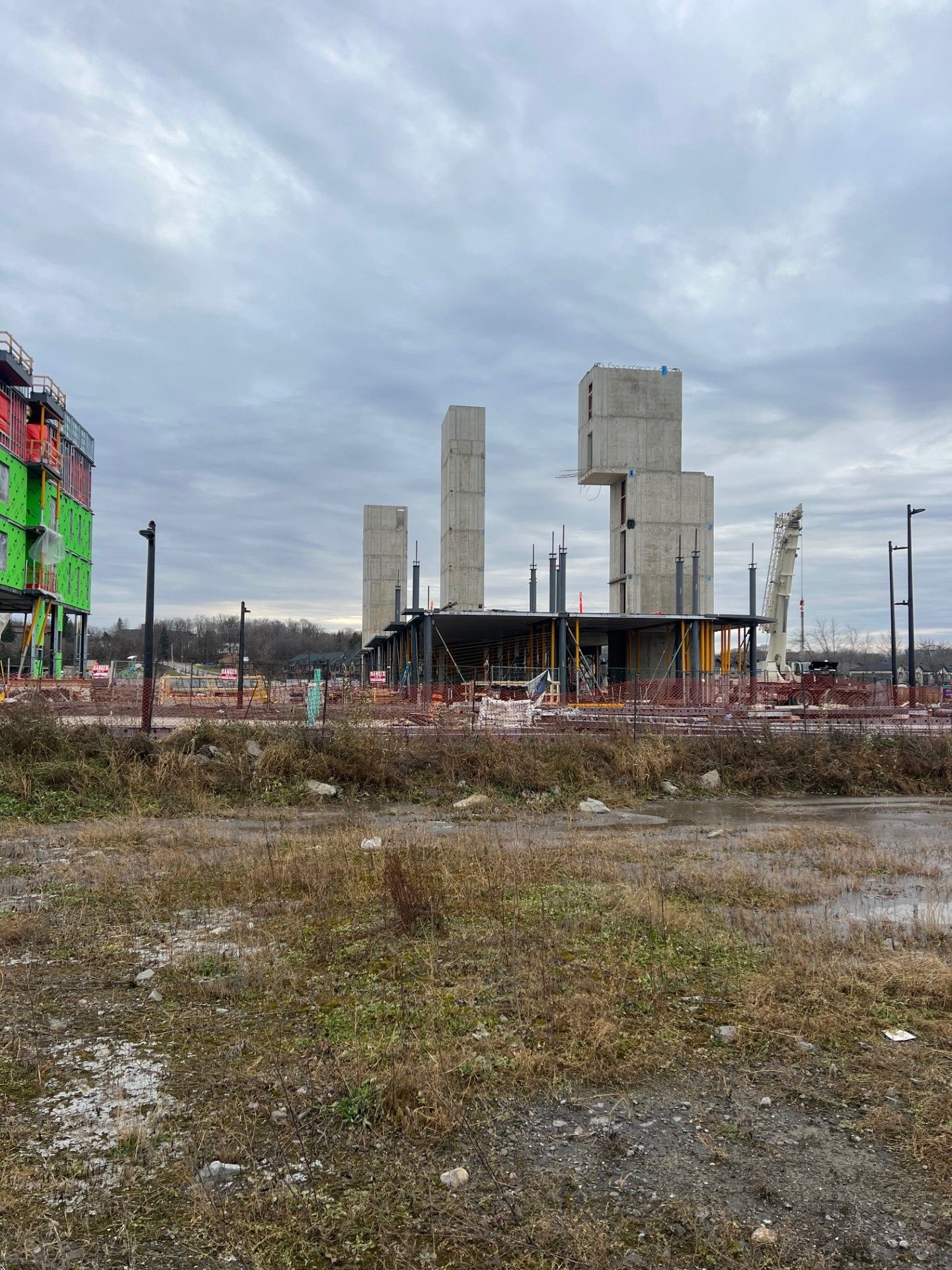 Construction site with tall concrete towers, a colorful building at left, and a cloudy sky over muddy ground.