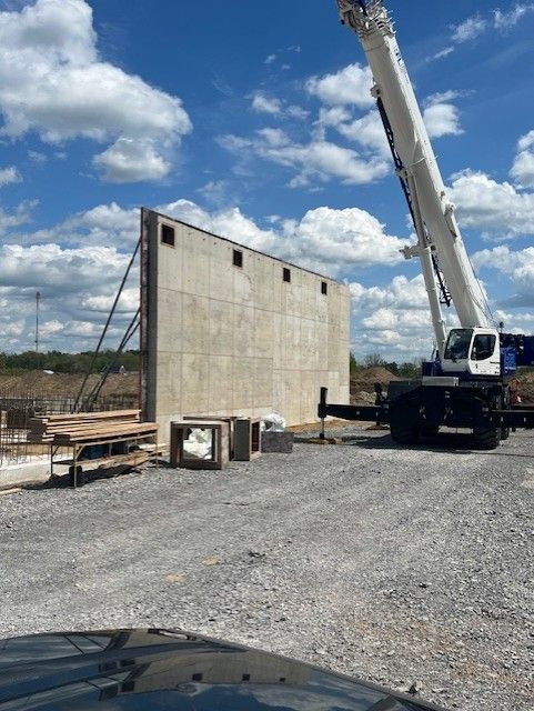 Construction site with crane lifting a concrete wall panel on a gravel lot under a partly cloudy sky