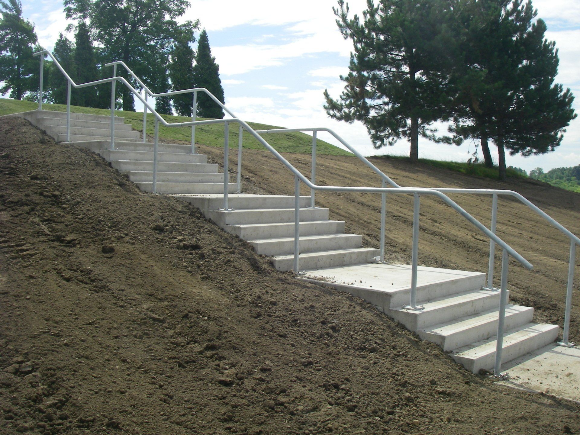 Outdoor concrete stairs with metal railings on a grassy hillside under trees and a partly cloudy sky