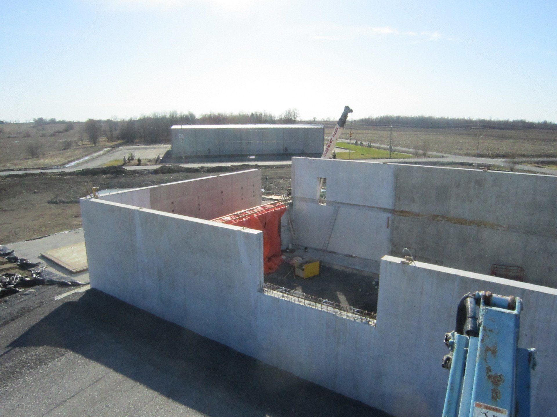 Concrete building foundation under construction in a flat rural area, with a blue excavator arm in the foreground.