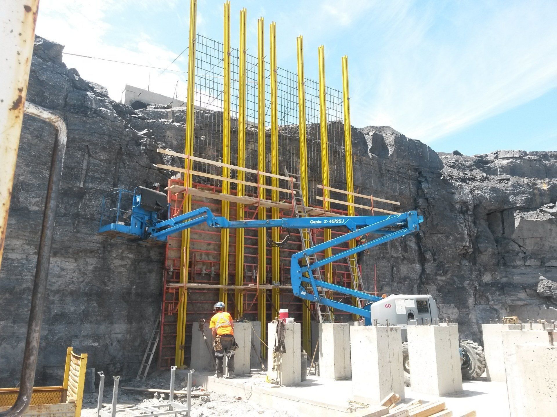 Construction site with workers beside blue concrete pump arms and tall yellow rebar against a rocky wall