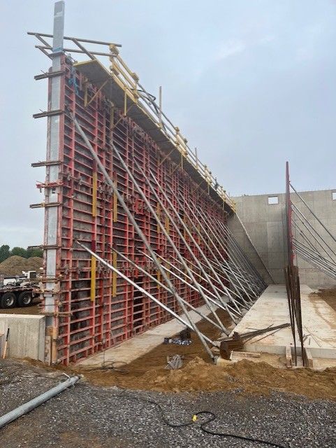 Tall concrete wall under construction with red steel scaffolding and support braces on a gravel site