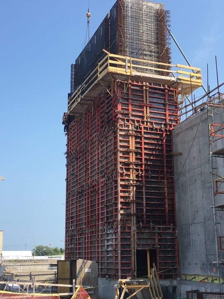 Tall concrete structure under construction with red scaffolding and wooden formwork against blue sky