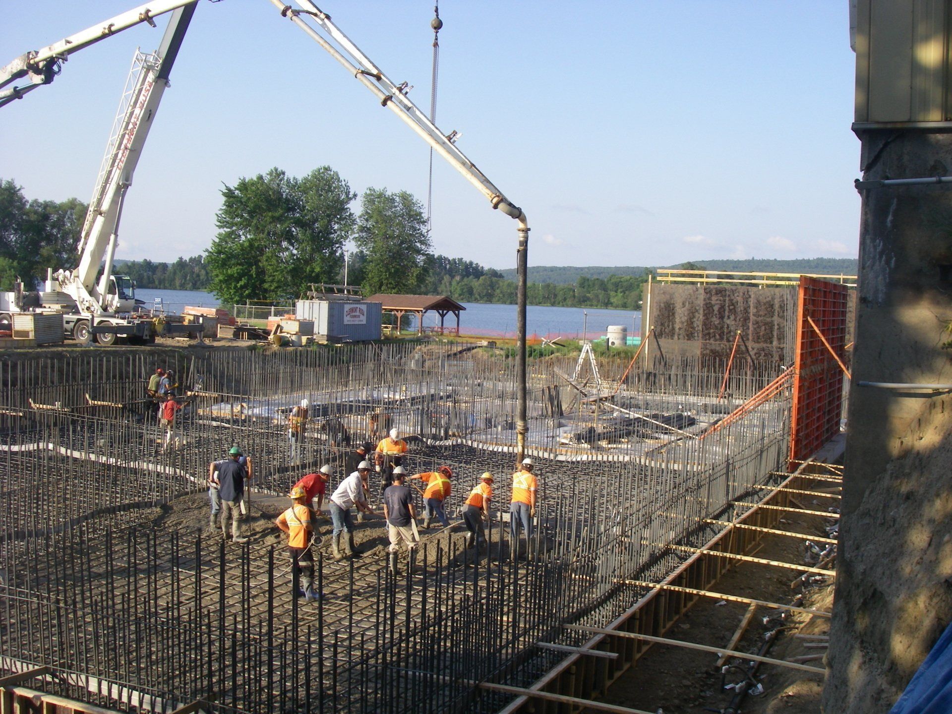 Workers pour concrete at a large construction site with rebar, cranes, and a lake in the background.