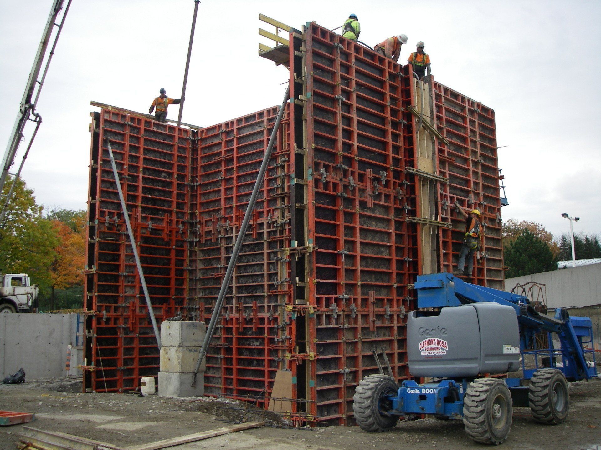 Workers atop a large red formwork structure, with a blue boom lift in a construction site.