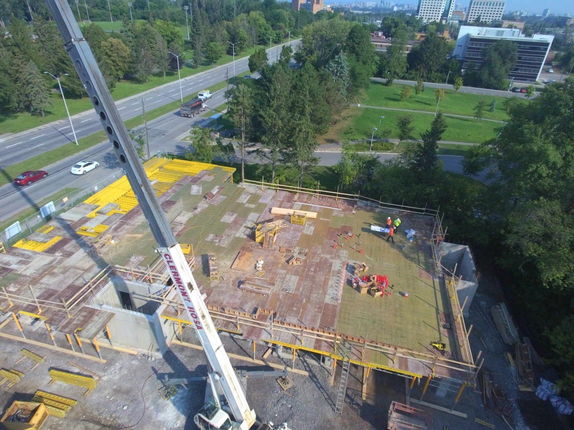 Construction site with a crane and workers on a concrete foundation beside a road and trees.