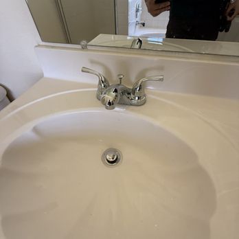White bathroom sink with chrome faucet, drain, and countertop, with a partially visible mirror and person reflected above.