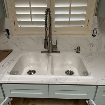 A kitchen sink with a high-arc faucet sits in front of a window with white shutters. Light blue cabinets are below.