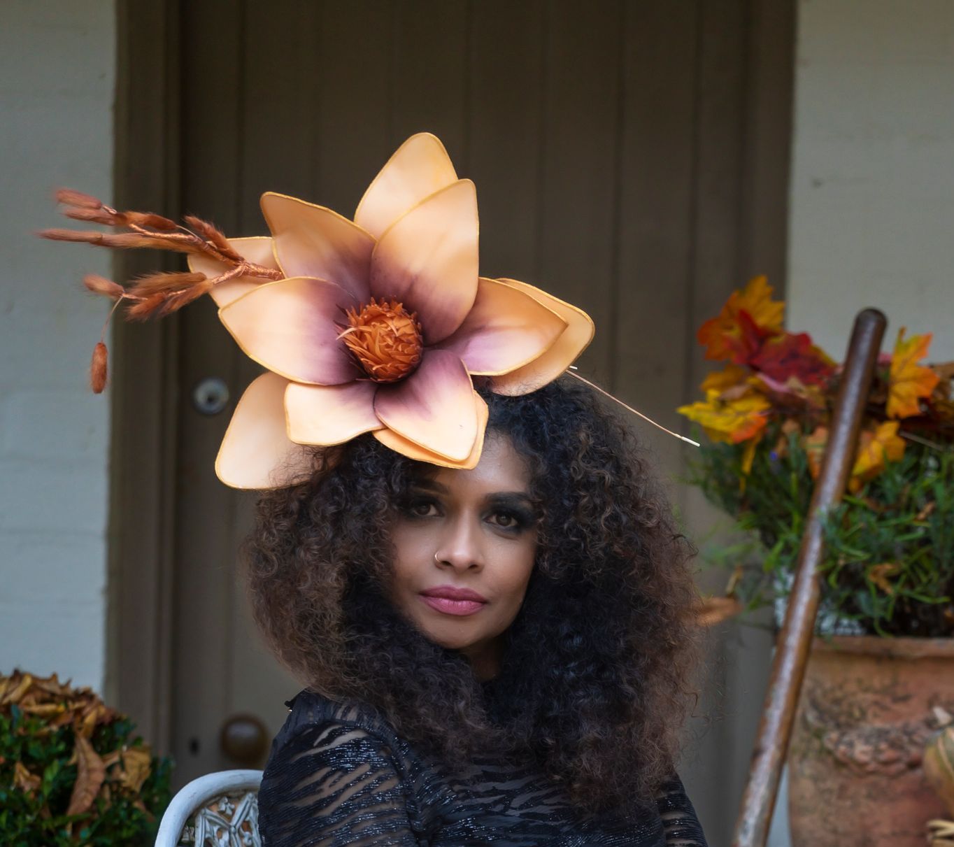 Woman in a large flower hat, standing outdoors beside potted plants and a wooden door.