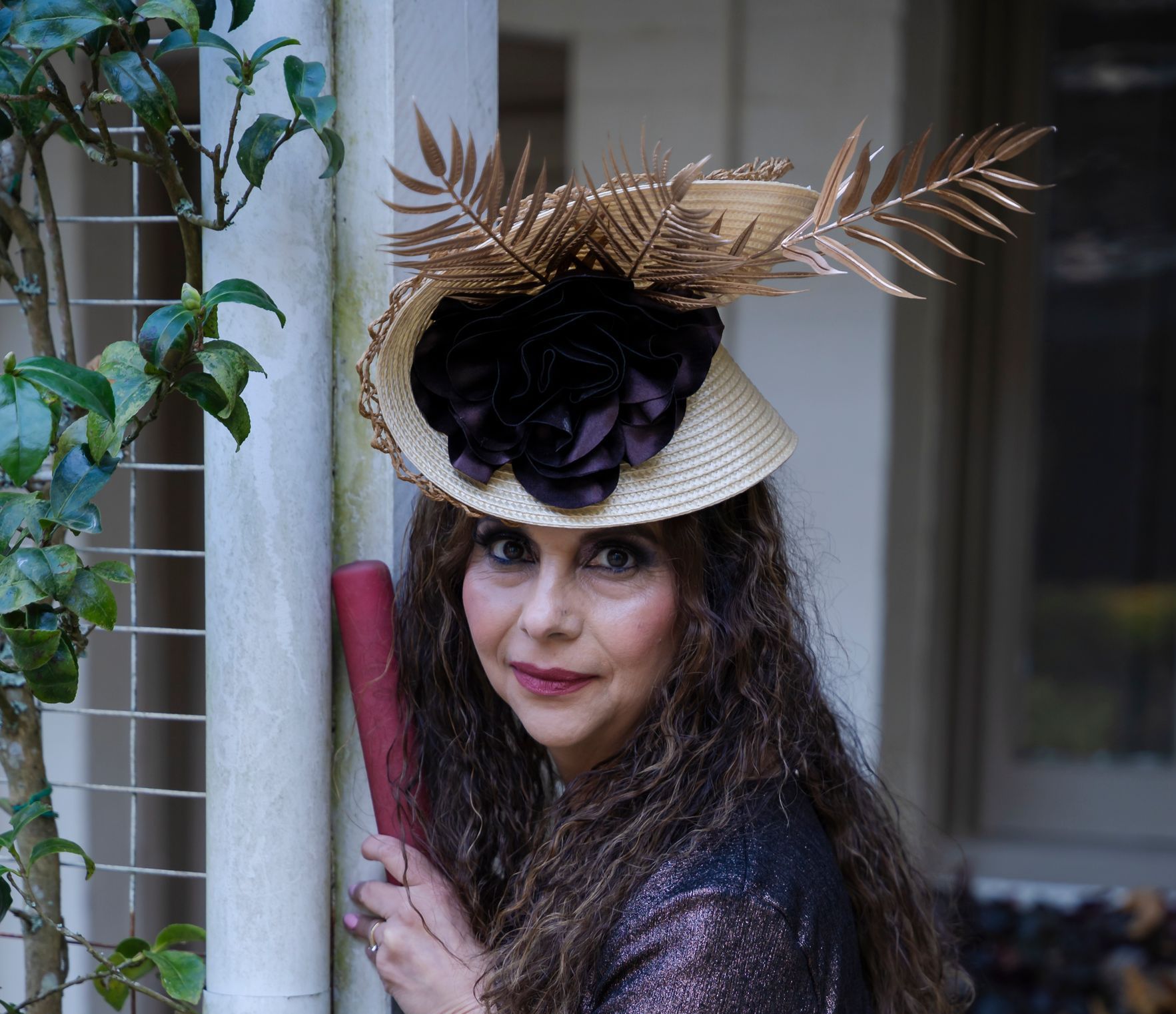 Woman in a decorative hat with black flowers and gold leaves, standing by a doorway with a pink cane.