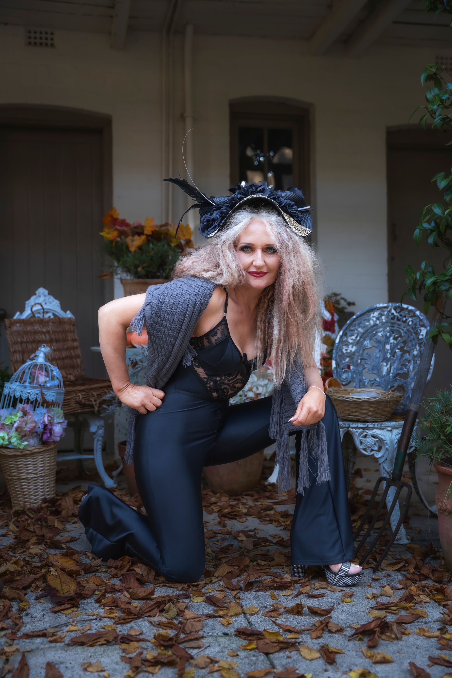 Person in a black outfit crouches on a porch amid autumn leaves, wearing a dramatic feathered headpiece.