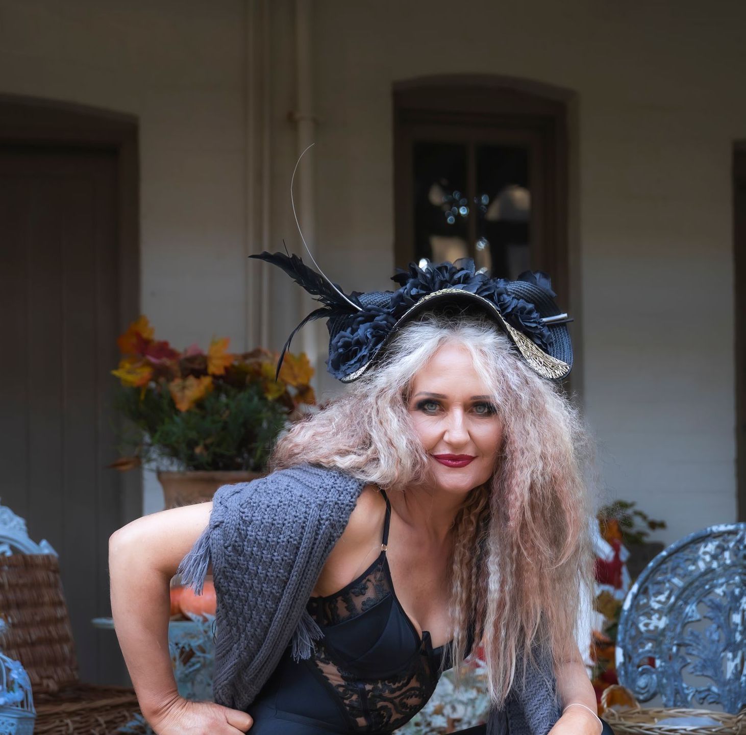 Woman in a black lace outfit and feathered hat posing on a patio with flowers behind her