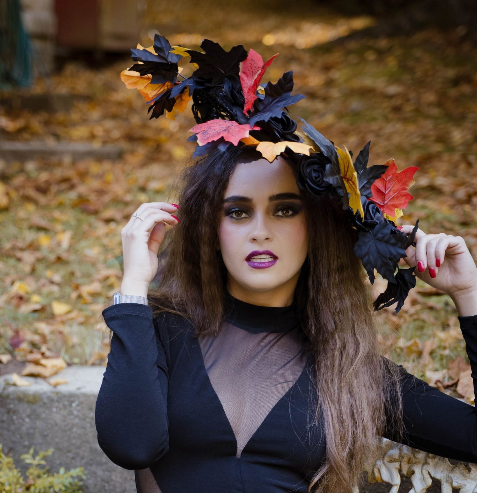 Woman in a black outfit wearing a leaf crown, posing outdoors with autumn foliage behind her