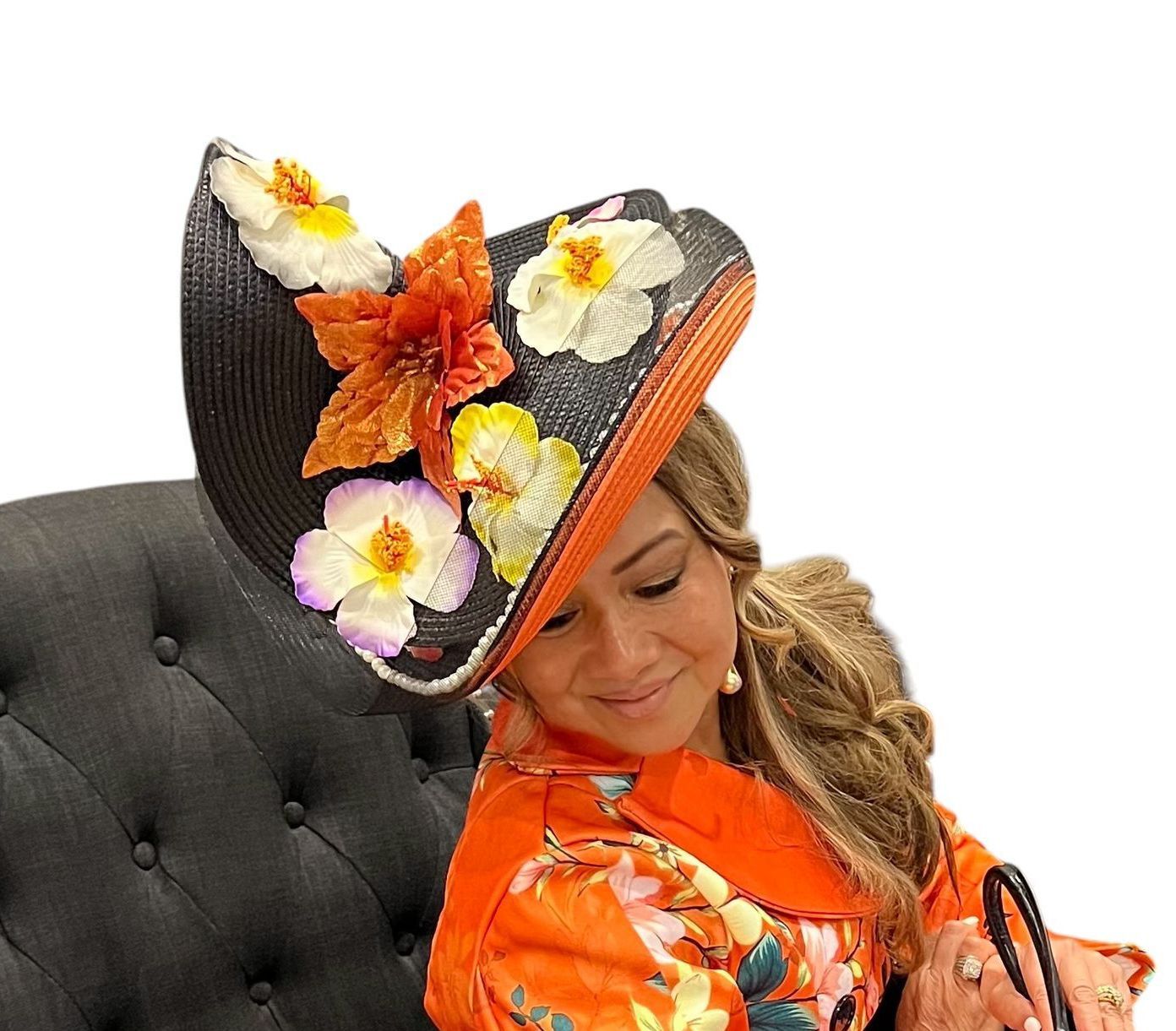Woman in orange outfit wearing a large floral hat, smiling with head tilted against a tufted black chair.