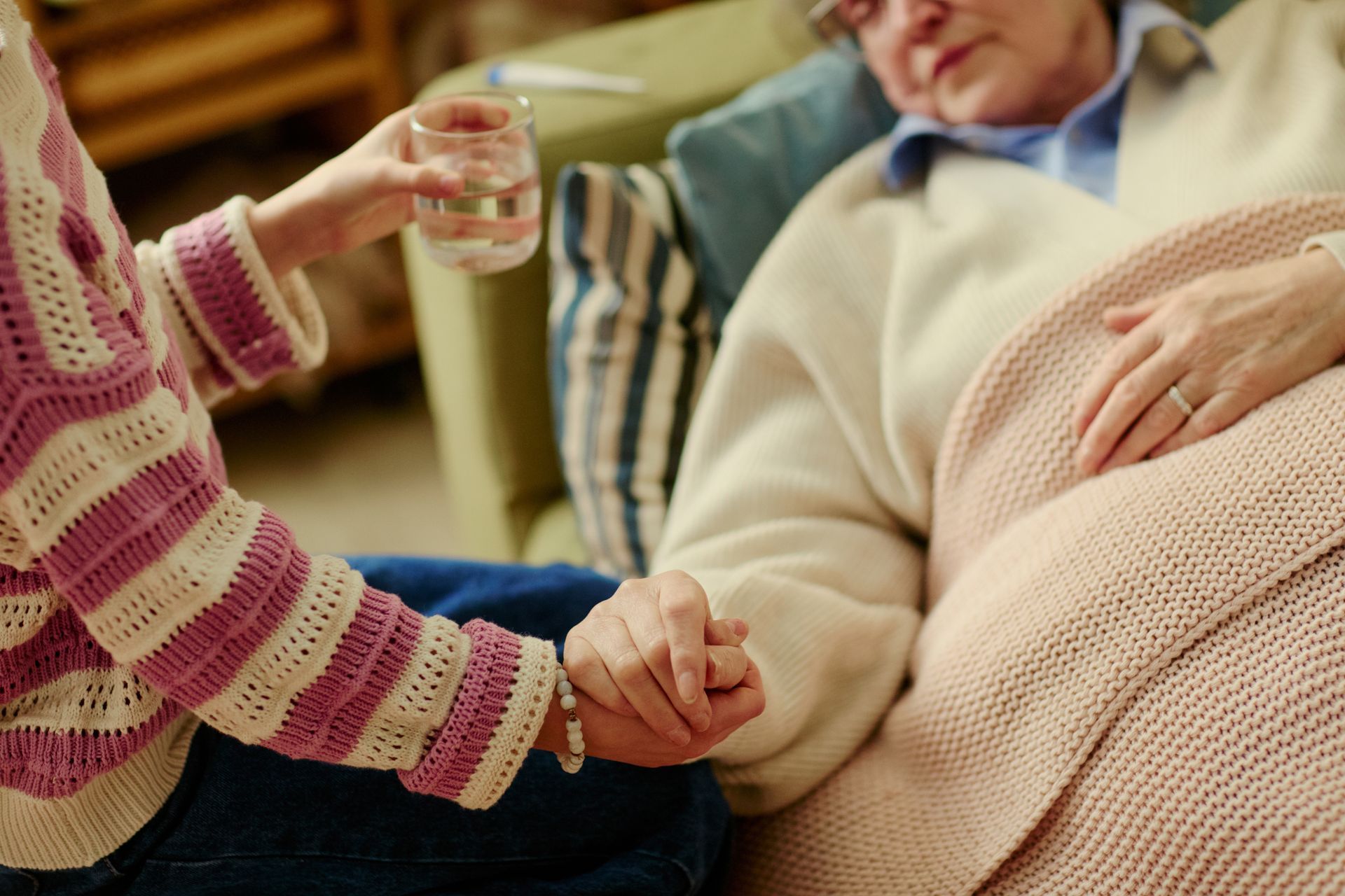 Person holding a resting patient’s hand beside a reclining figure with a glass of water