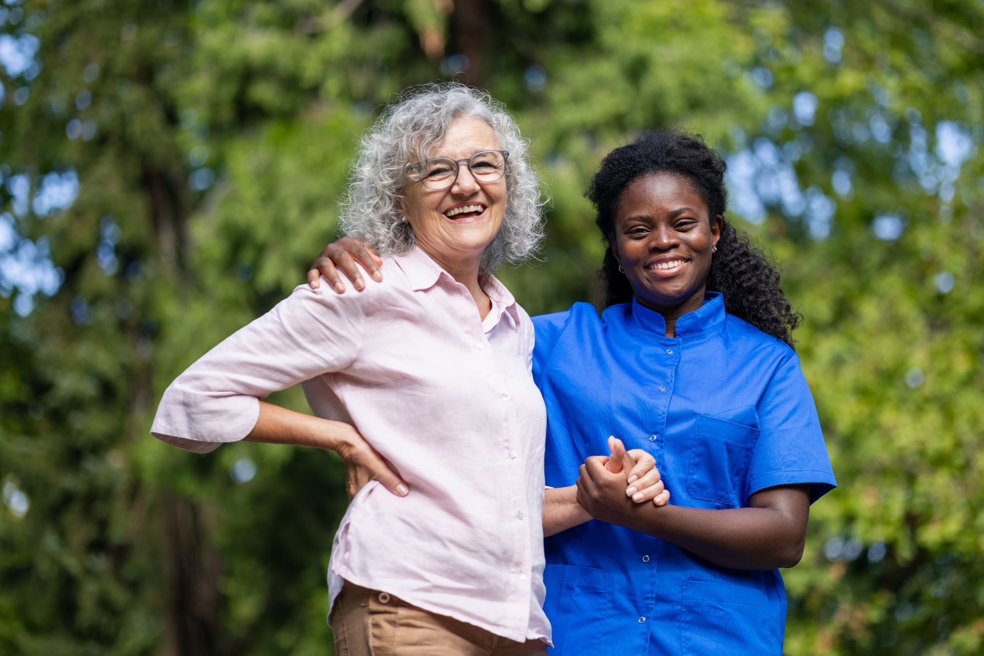 Two people smiling outdoors, one in a light pink shirt and one in a blue shirt, standing in a green park.