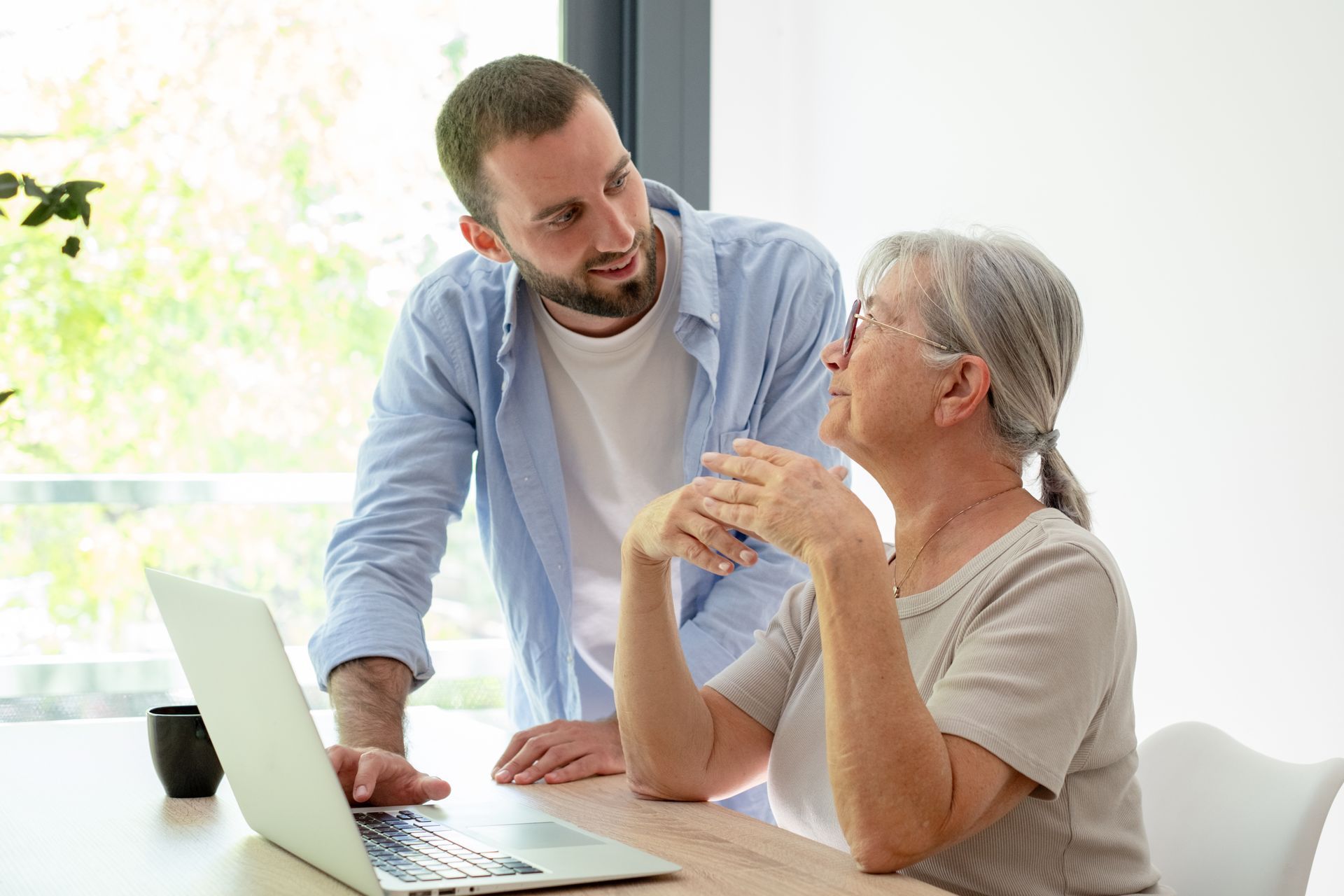 Man showing a laptop to an older woman at a bright table, both engaged in conversation