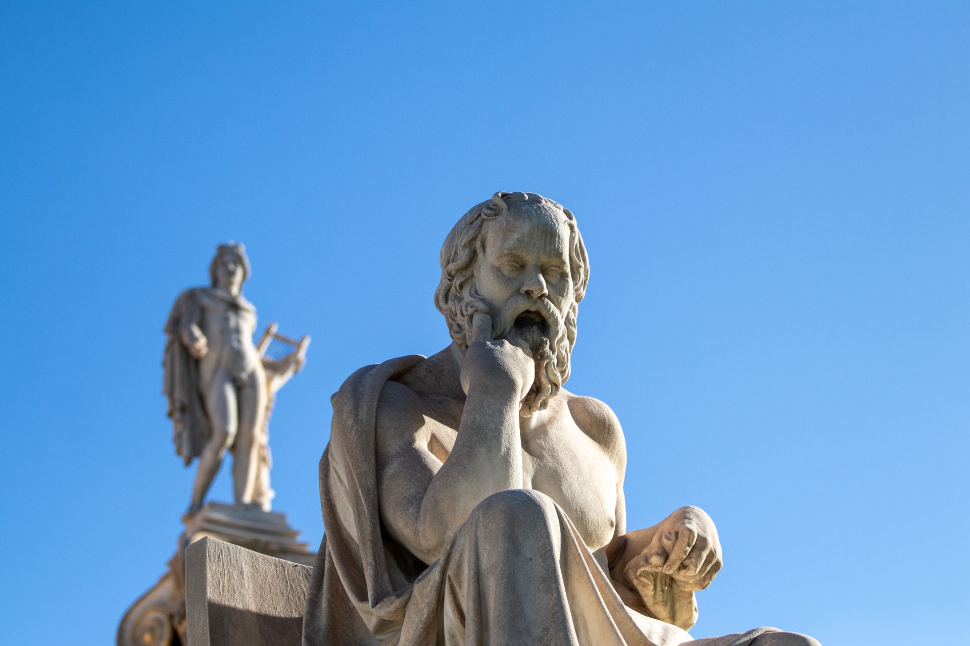 Two stone statues against a clear blue sky, one seated in the foreground and one standing behind.