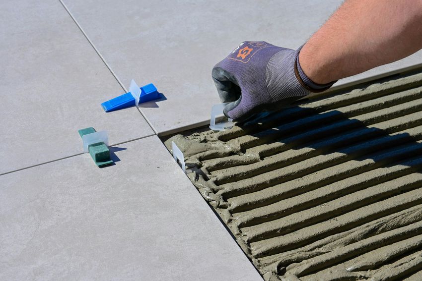 Gloved hand spreading mortar on a tiled floor beside spacers and a trowel-marked surface
