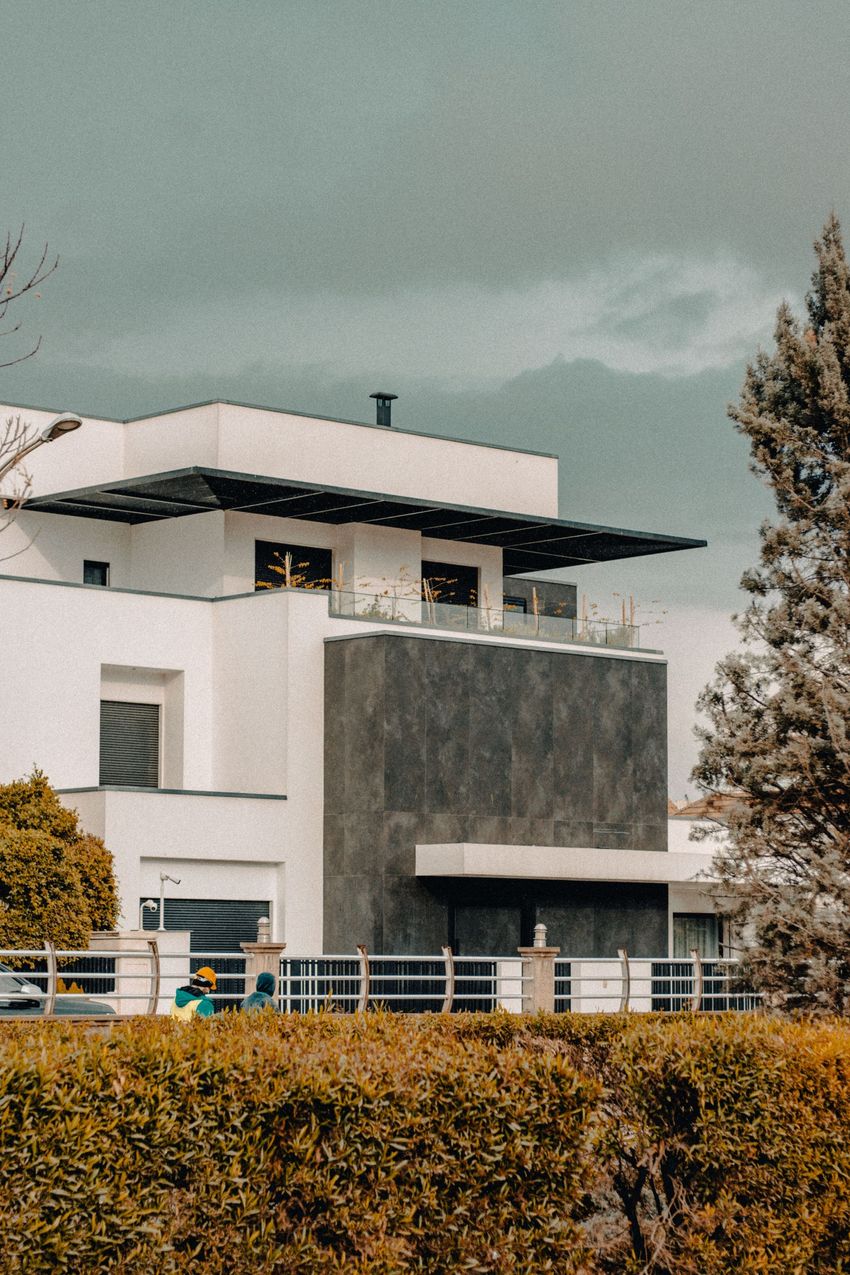 Modern white house with a dark balcony, hedge in front, and cloudy sky above