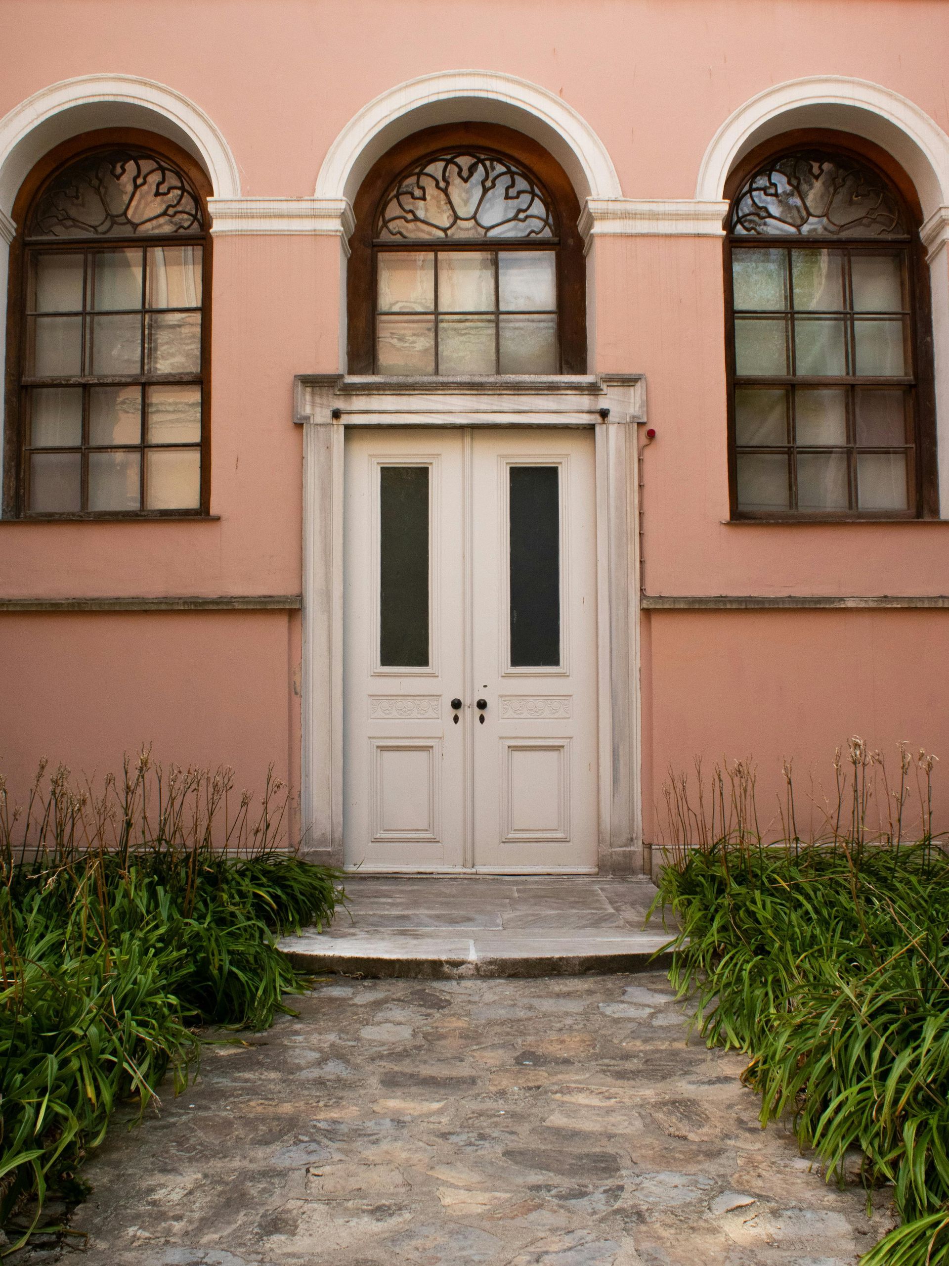 Pink building facade with white double door, three arched windows, and a stone walkway bordered by greenery