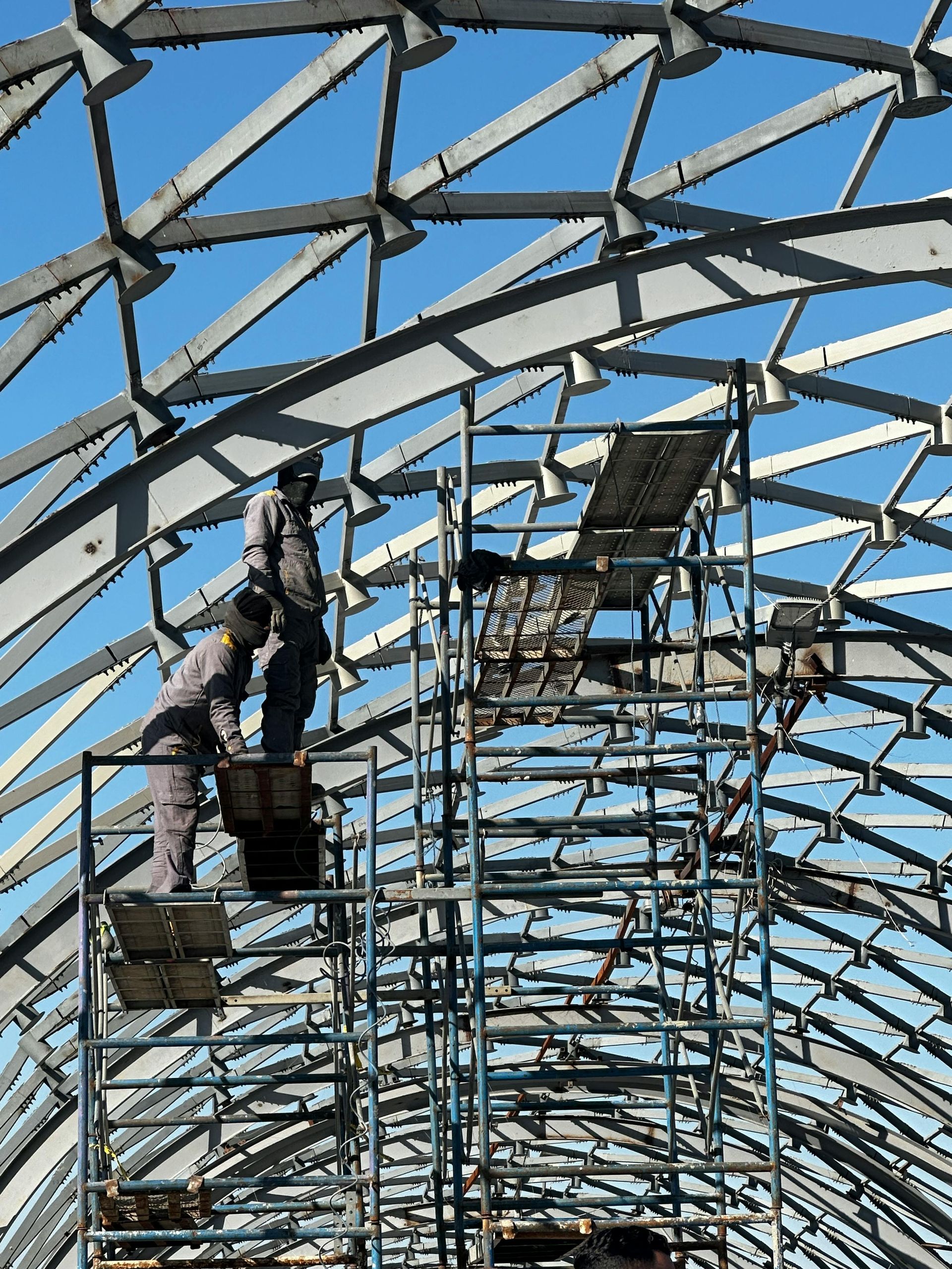 Workers on scaffolding inside a large arched steel frame against a blue sky